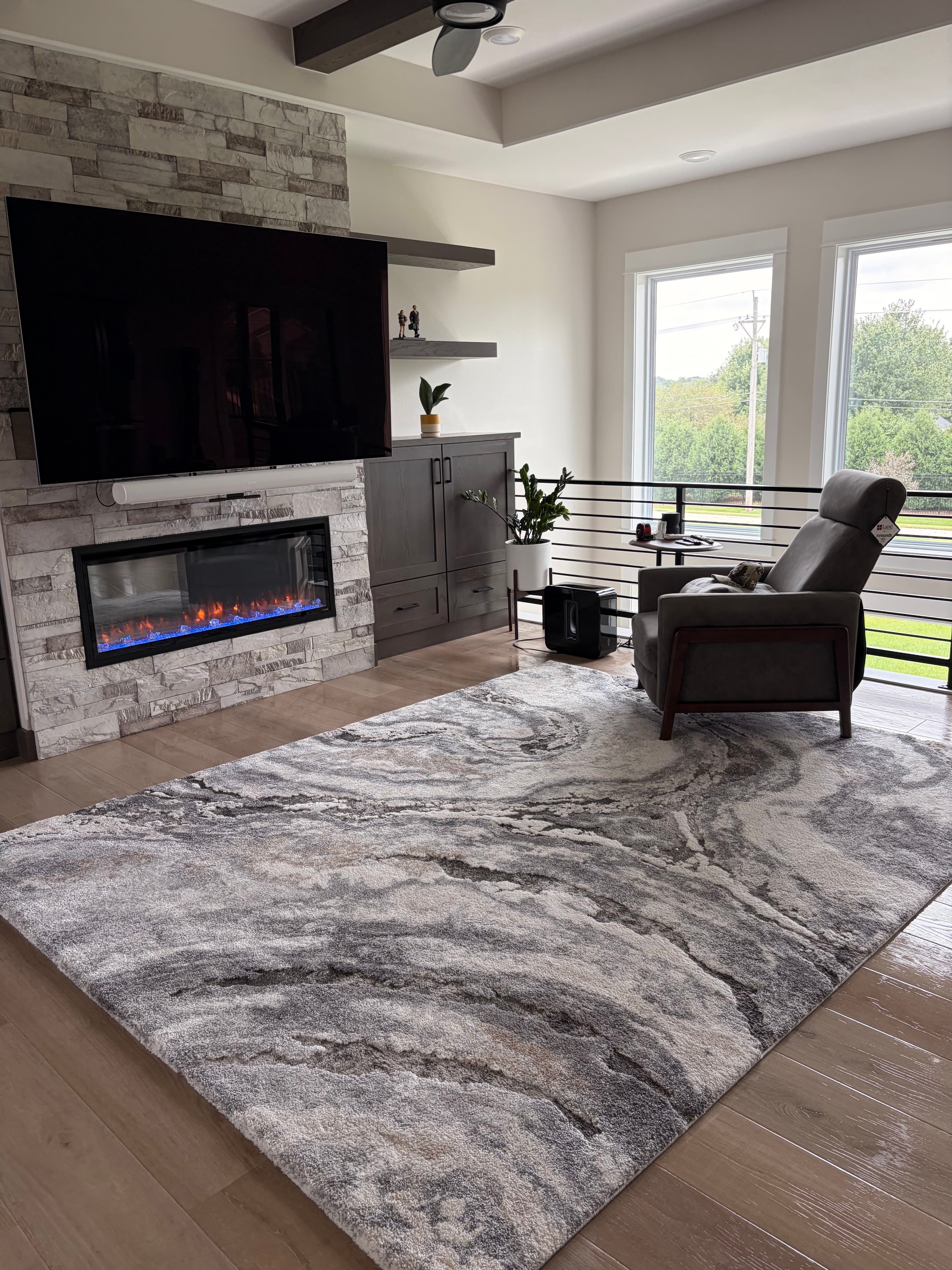 Modern living room with stone fireplace, wall-mounted TV, marbled rug, and grey recliner.
